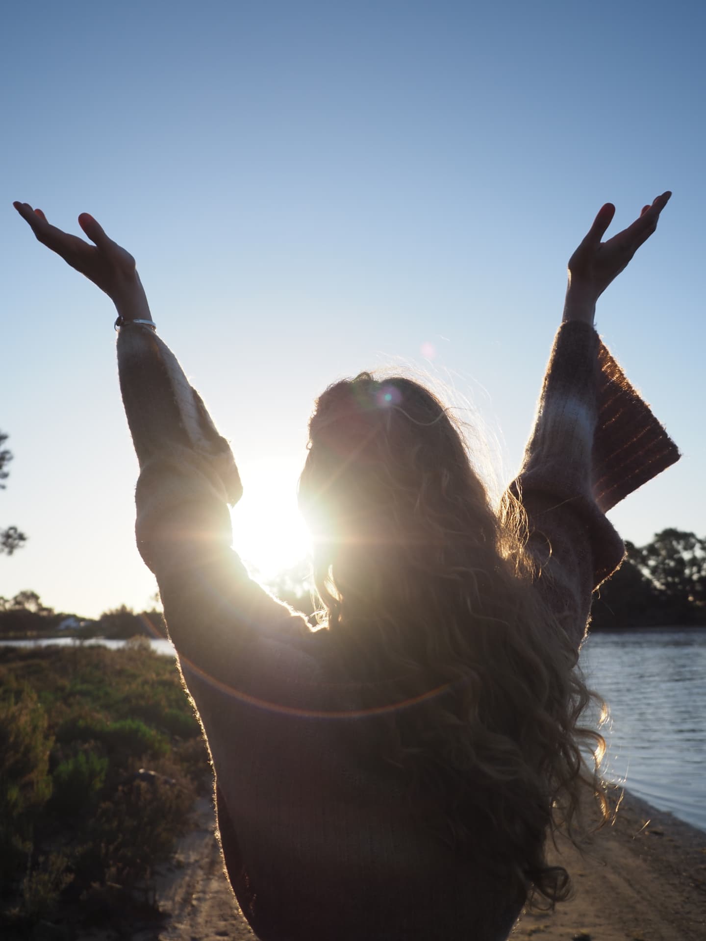 Anna-Lena seen from behind, arms raised openly toward the low sun over the water